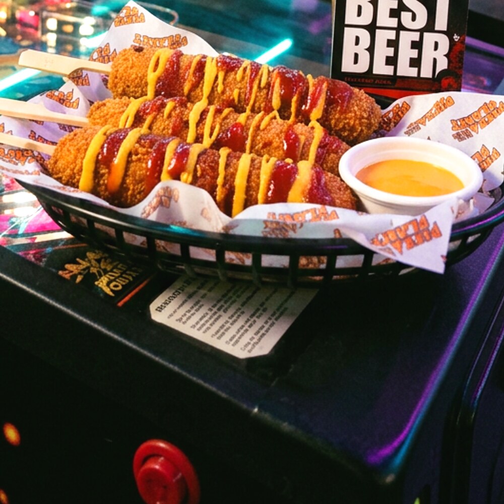 Three Planet Corndogs in a black basket with ketchup and mustard drizzle and a pot of beer cheese sauce, on a pinball machine at Planet Royale.