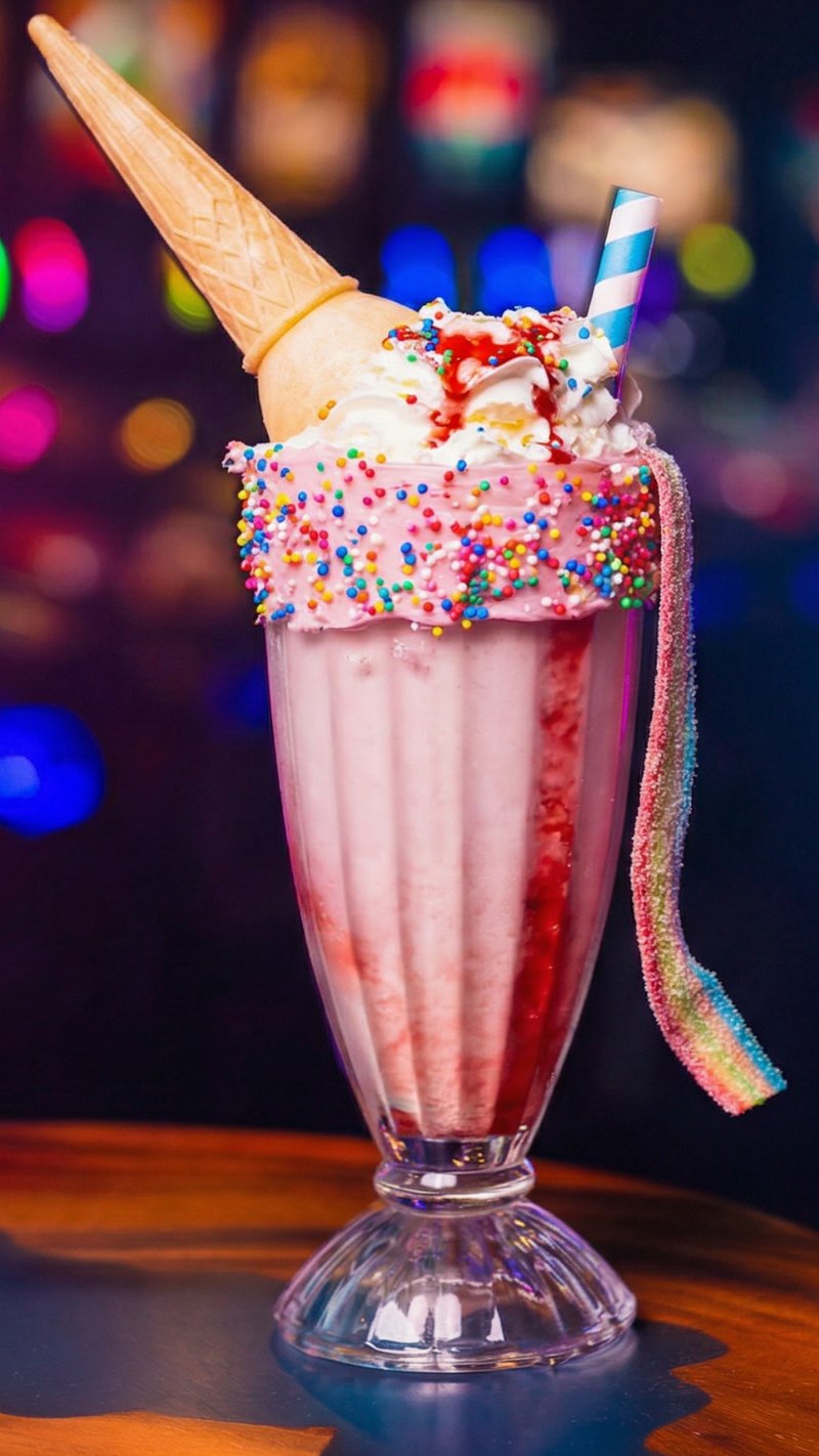 Vanilla Unicorn shake - pink strawberry shake with rainbow sprinkle rim, sour rainbow strap, ice-cream cone topper and whipped cream against arcade neon.
